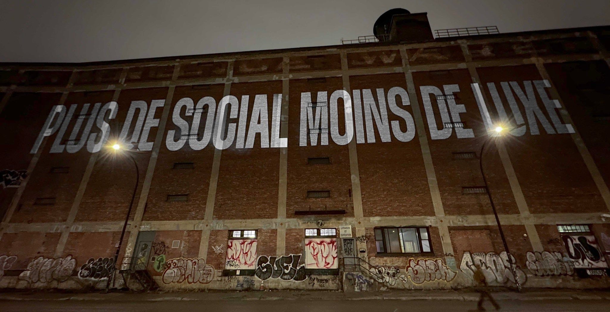 Projection sur l’entrepôt Van Horne, la nuit. On peut y lire: « PLUS DE SOCIAL MOINS DE LUXE »