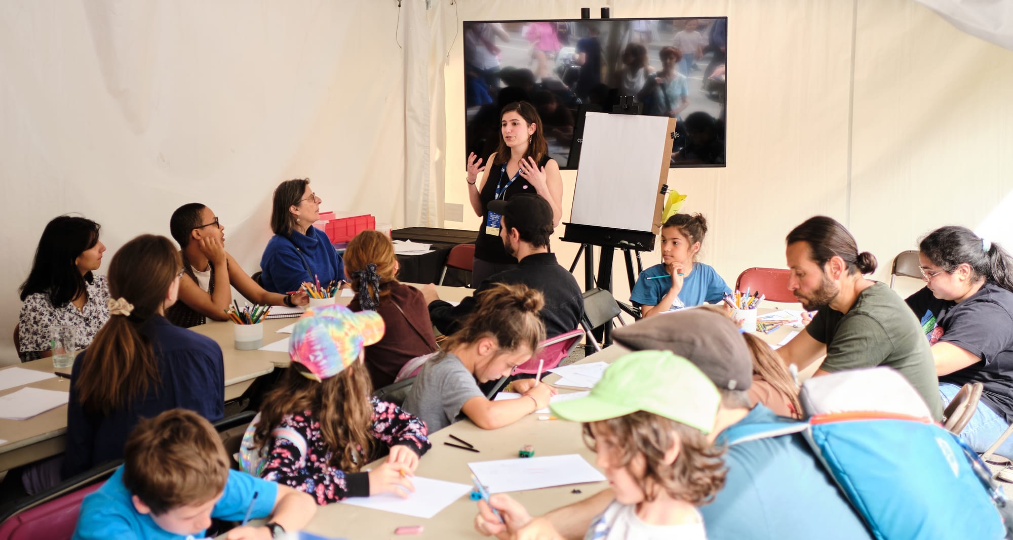 Une vingtaine de personnes attablée lors d'un atelier de dessin lors d'explications de l'animatrice de l'activité, debout.
