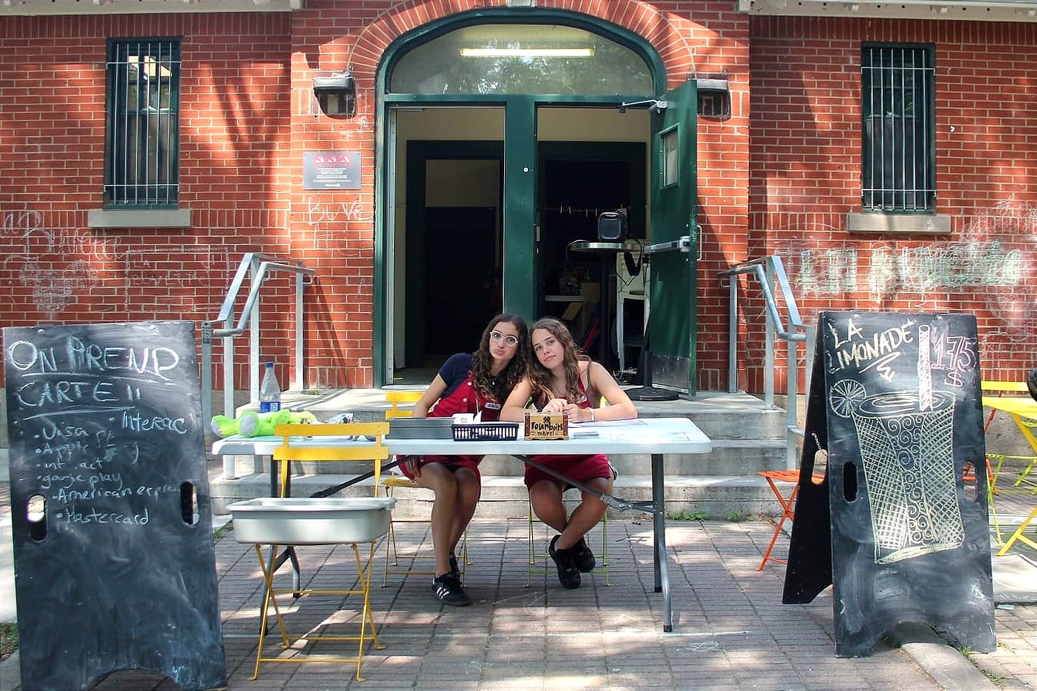 On voit les deux filles devant le chalet, avec des tableaux à leurs côtés qui indiquent les modes de paiement et vantent la limonade à 1,75$.