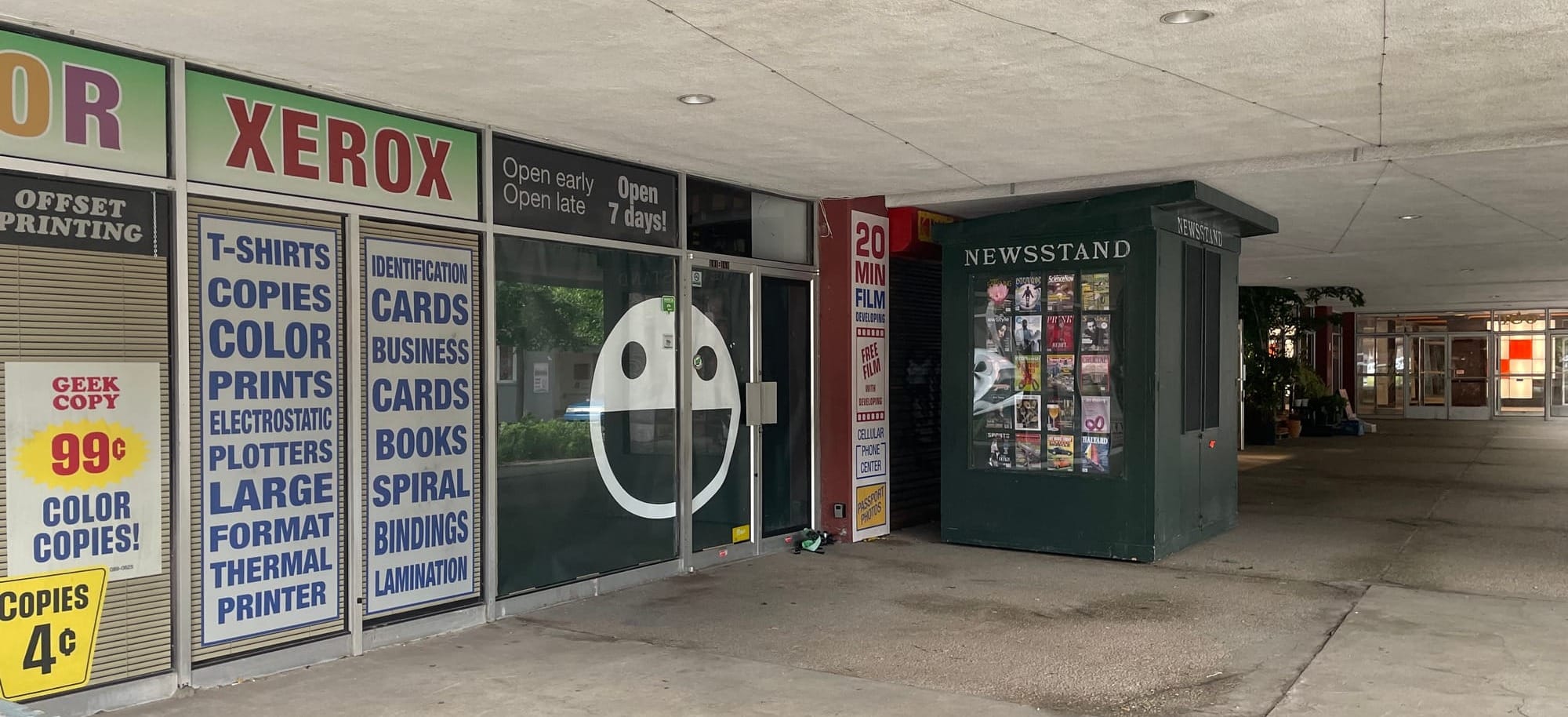 La vitrine d'un ancien restaurant est couverte d'affiches en anglais qui présente une imprimerie et l'on voit un ancien kiosque à journaux.