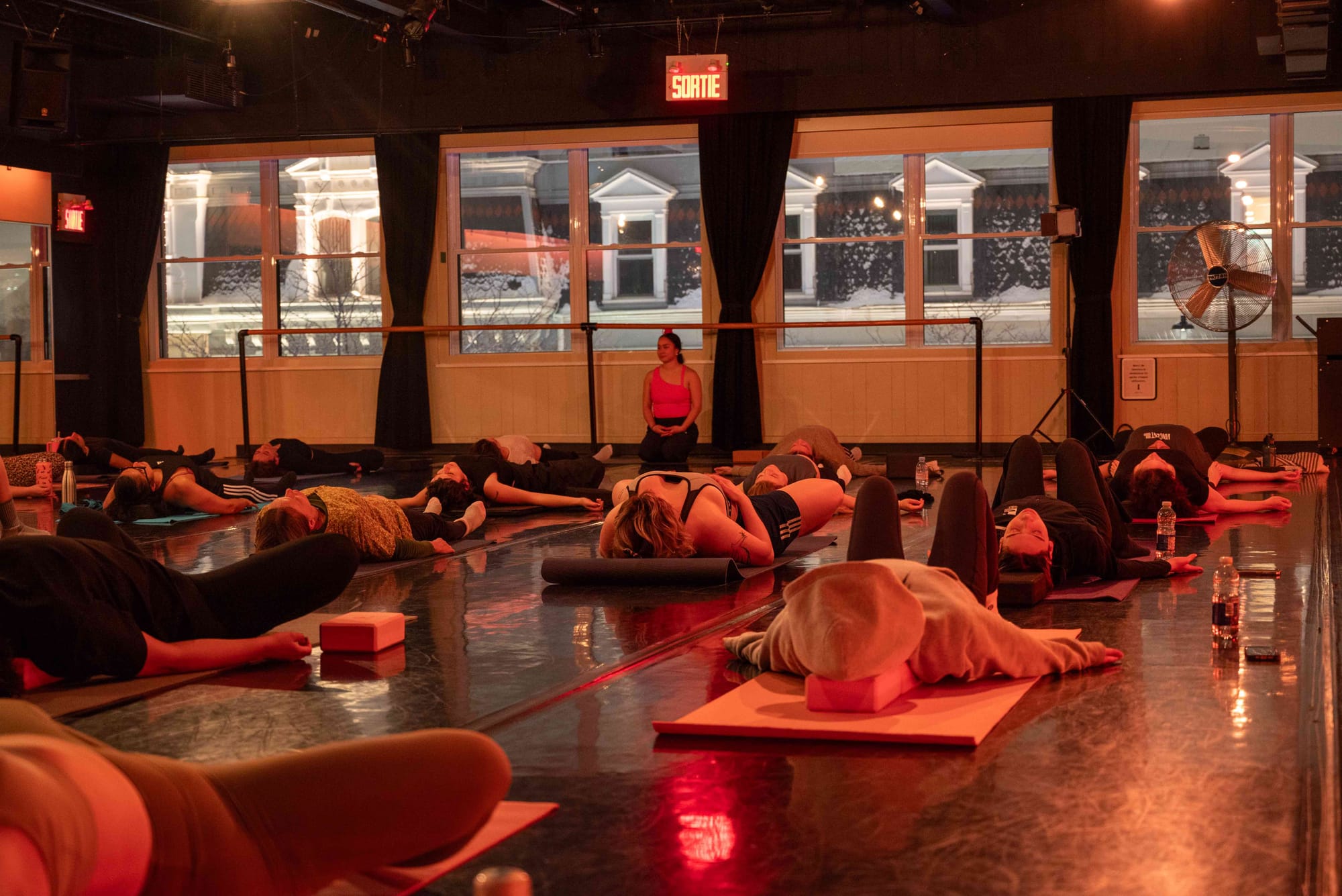 Une des grandes salles du Studio Bizz, bordée de fenêtres, où une dizaine de personnes sont allongées sur des tapis de sol dans le cadre d'un cours.