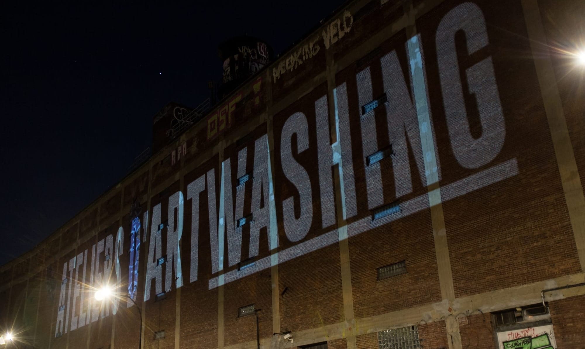Les mots "ATELIERS D'ARTWASHING" sont projetés en énores lettres majuscules sur la façade de l'entrepôt Van Horne, la nuit.