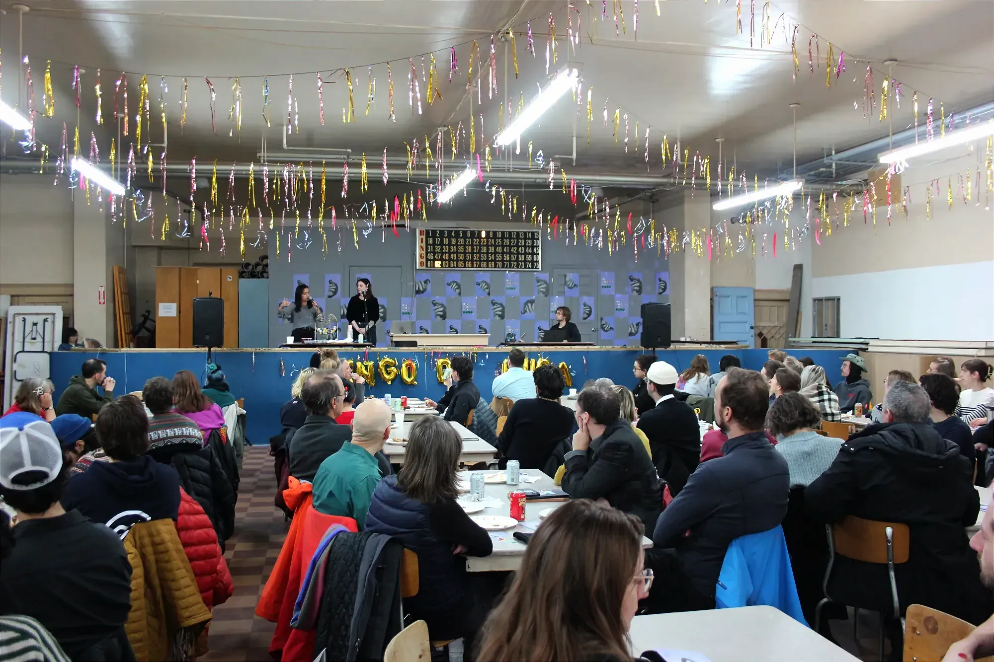 Vue du fond de la salle. On voit la foule et, au loin, Mme Ghazal parlant au micro, aux côtés de Coralie Laperrière.