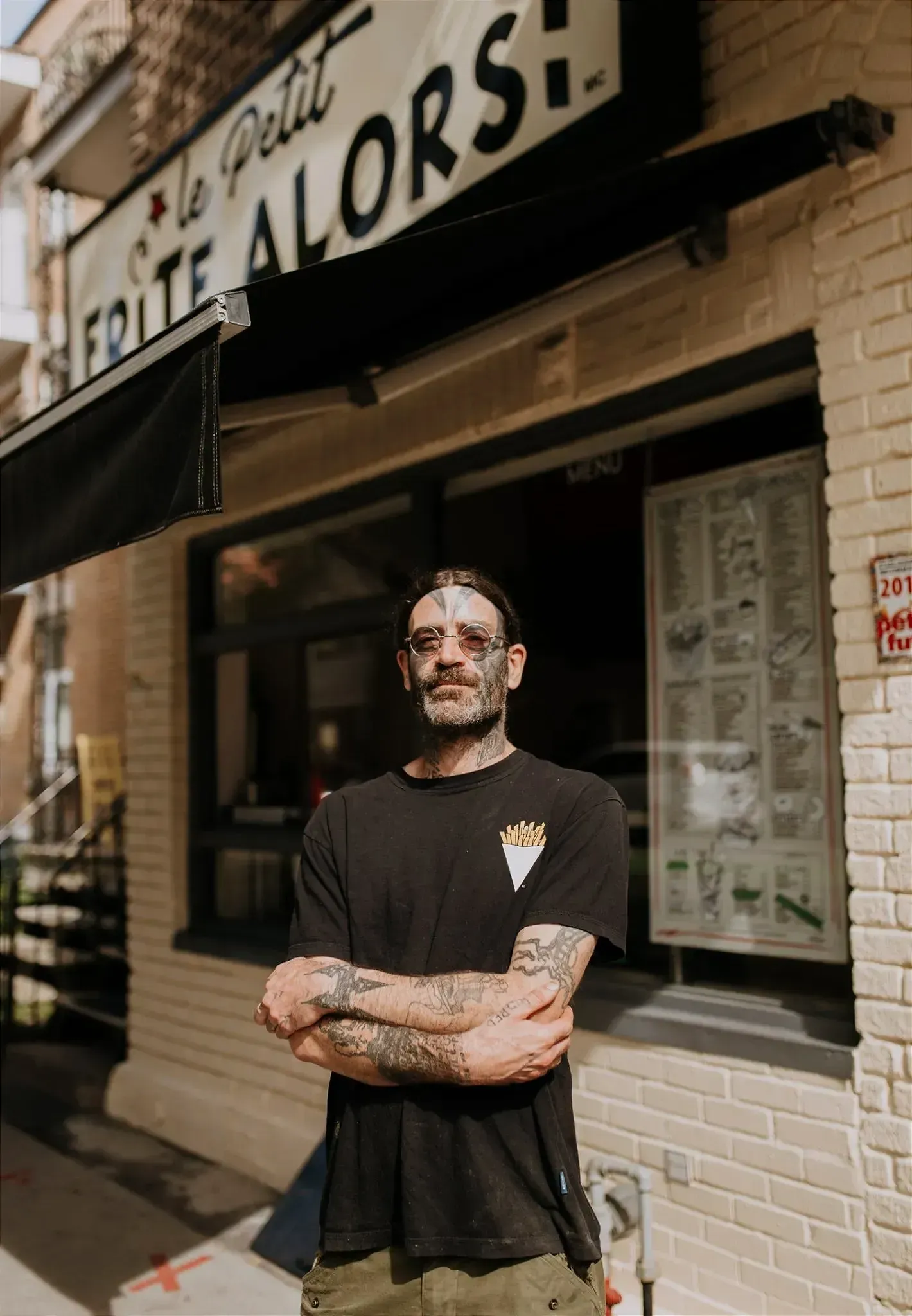 M. Saint-Jean, tatoué aux bras et au visage, pose devant la fenêtre du Frite Alors! sur Laurier.