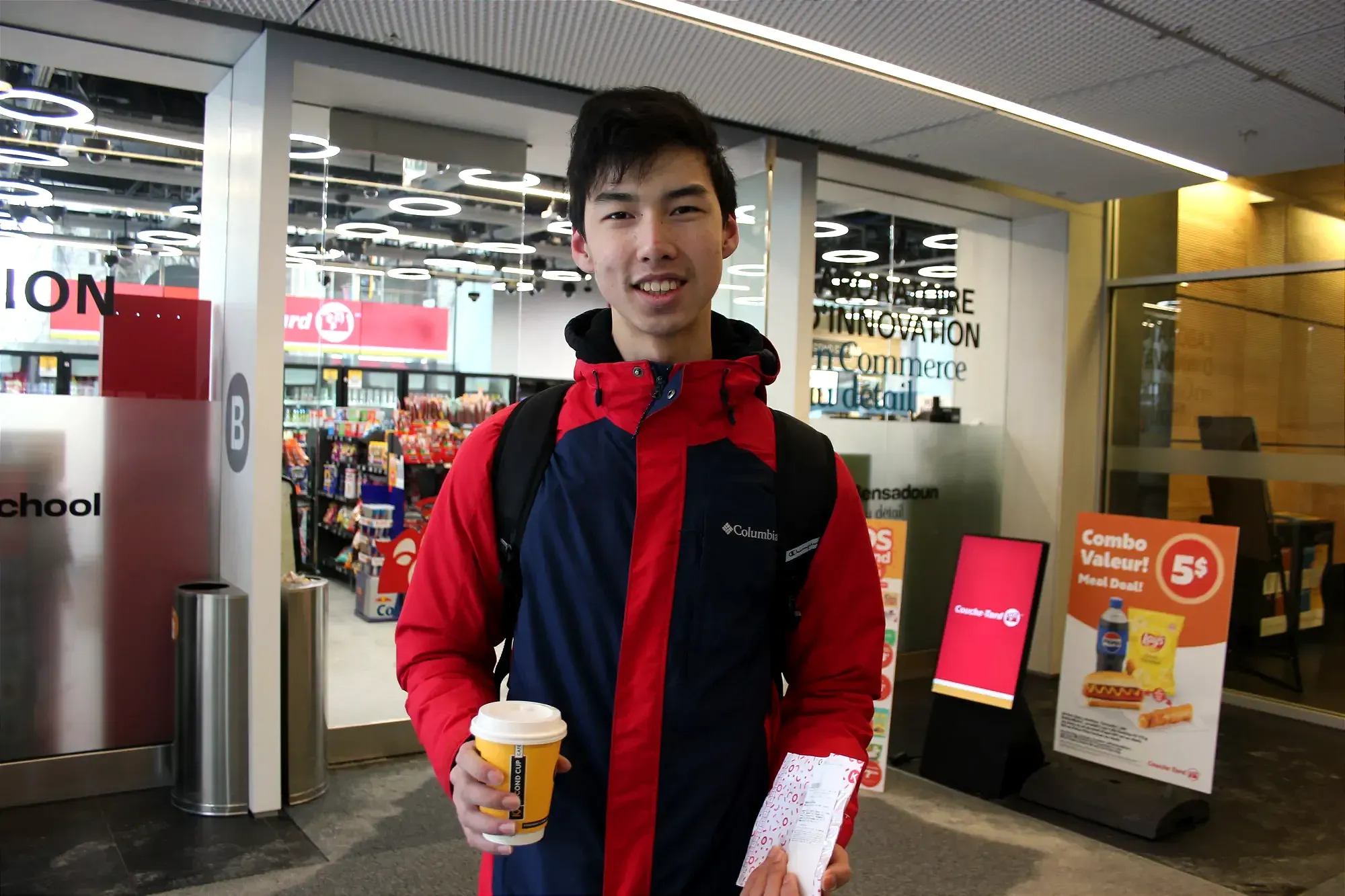 M. Bao pose devant l'entrée du dépanneur, tenant un café et une pâtisserie dans ses mains. 