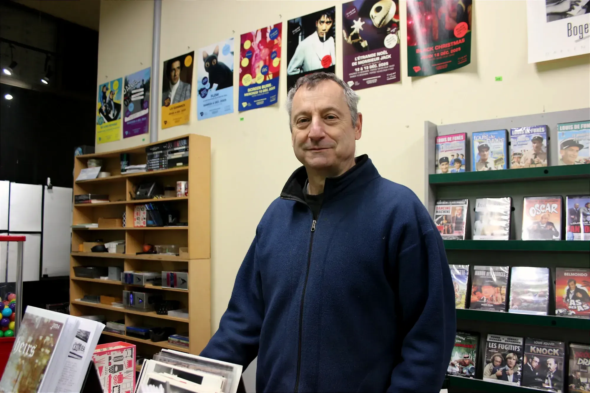 M. Major pose derrière le comptoir. On peut voir les affiches de film de Cinéma en Cinoche derrière lui.
