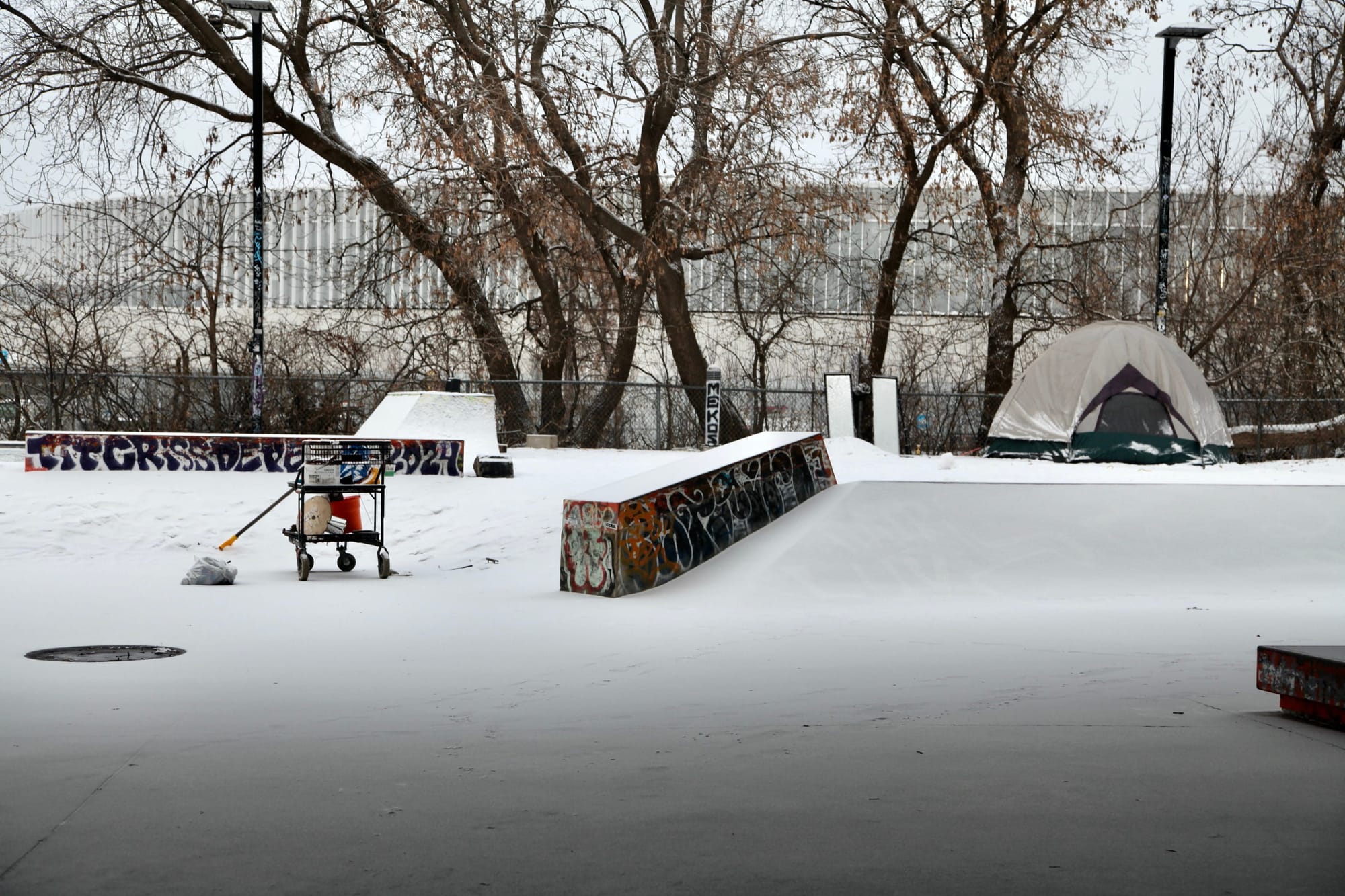 Une photo hivernale, avec de la neige au sol, une tente située près d'une voie ferrée et un chariot d'épicerie rempli d'objets.