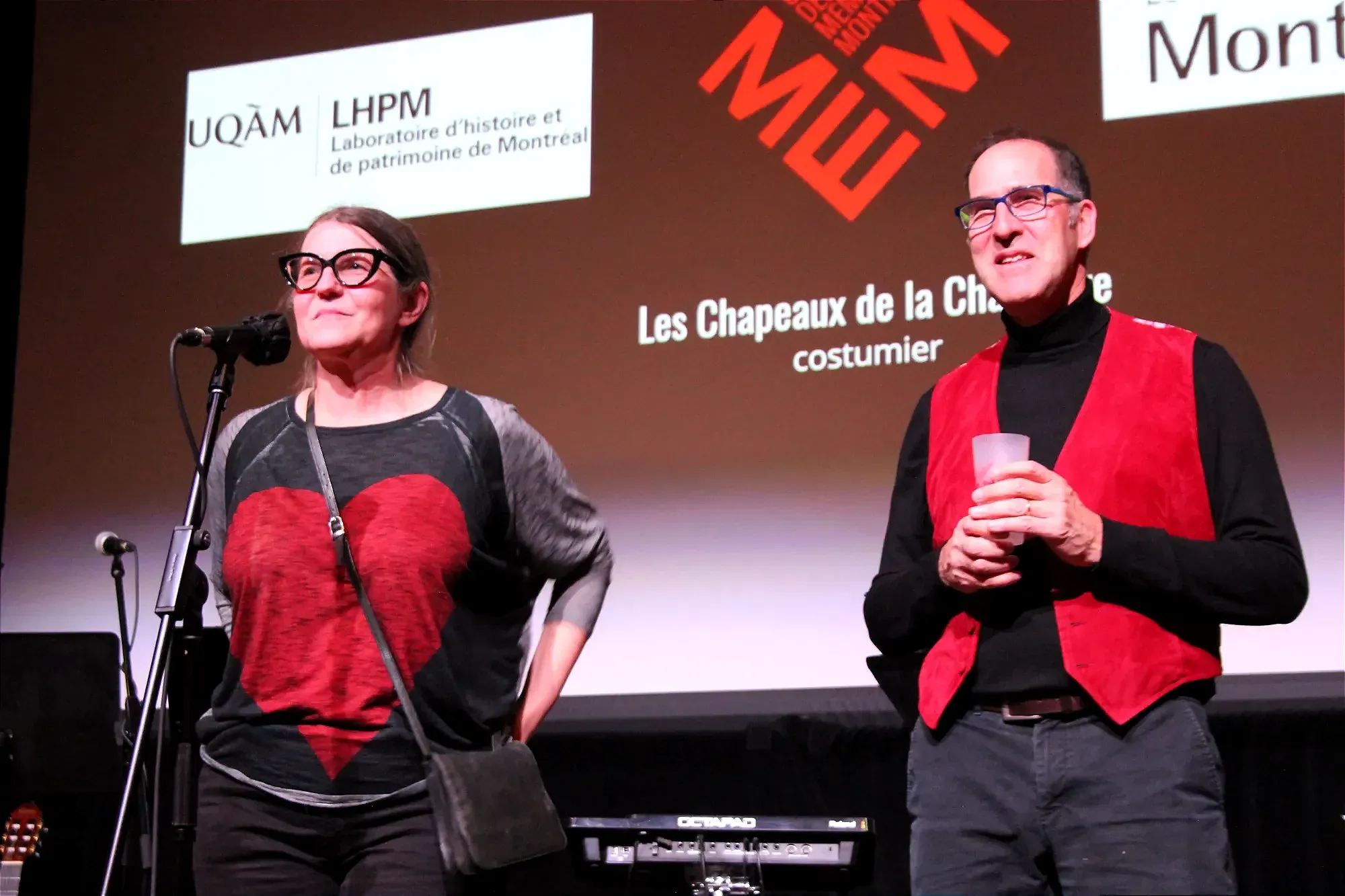 Mme Bandulet se tient derrière le micro, portant un chandail largement recouvert d'un grand cœur. M. Lacasse, porte une veste rouge et tient un breuvage dans ses mains.