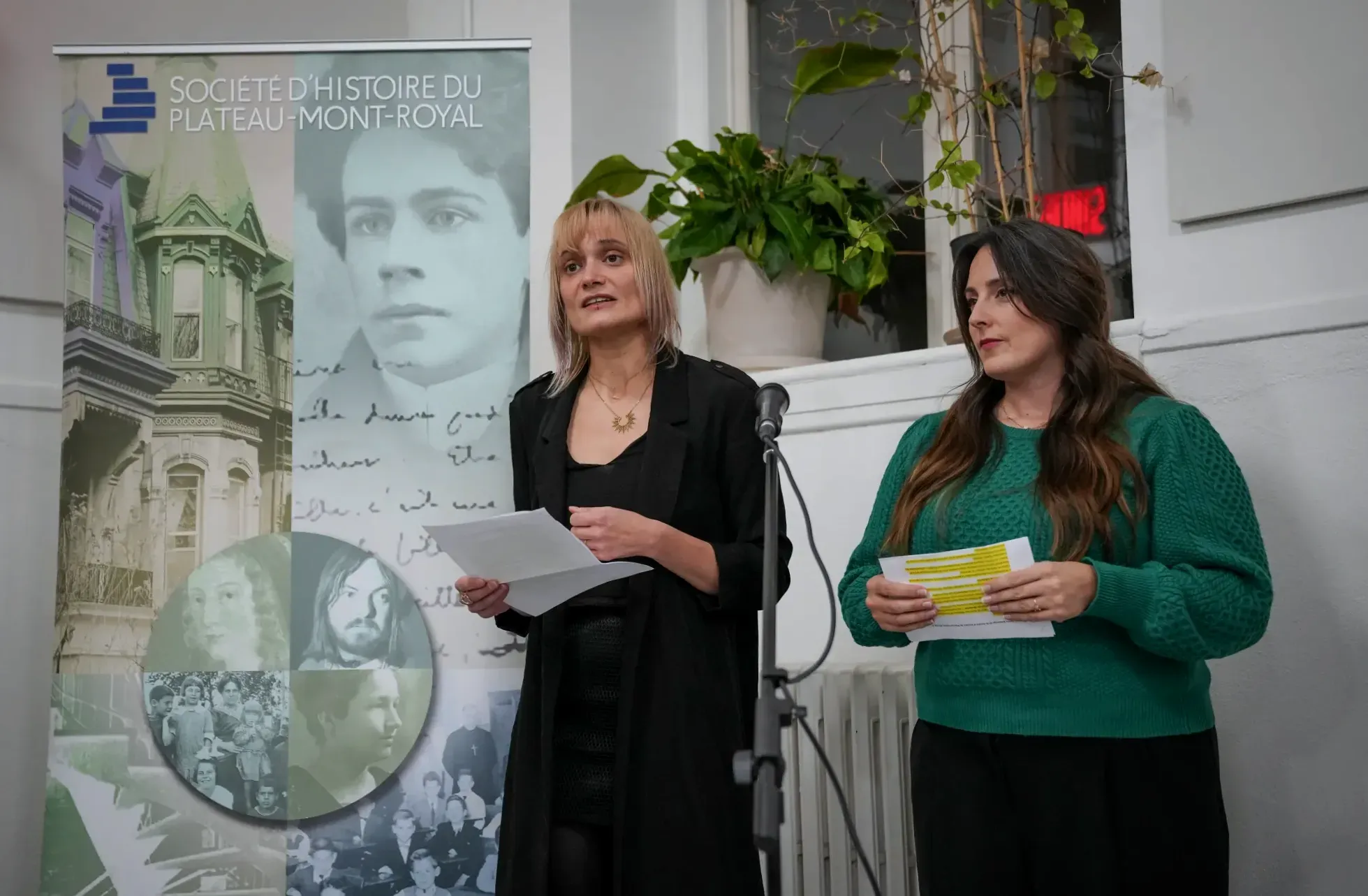 Mme Hüni prend la parole derrière un micro et devant une bannière de la Société d'histoire du Plateau, accompagnée de Marianne Rodrigue.