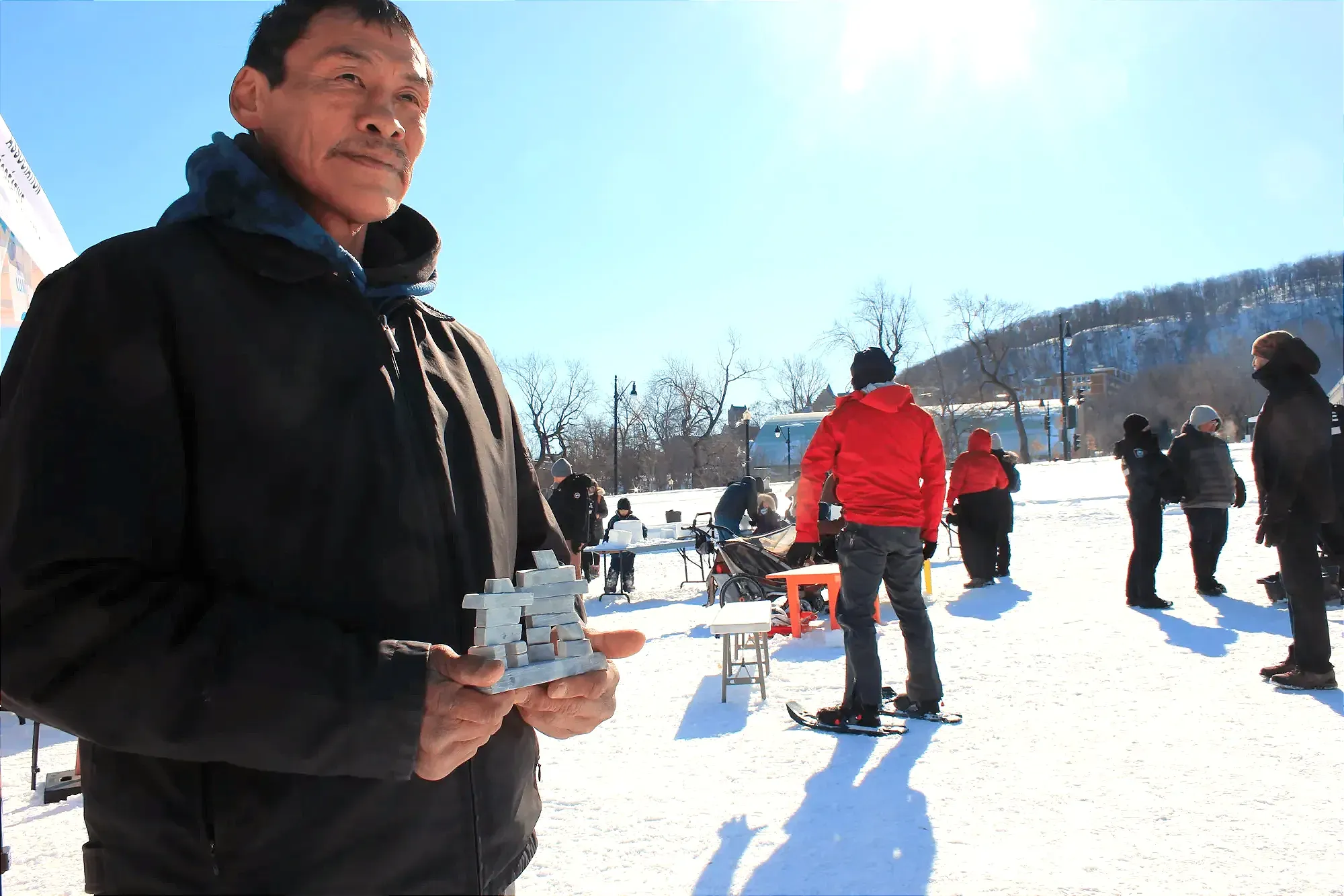 M. Komak tient son œuvre dans ses mains, regardant au loin, alors que l'on aperçoit des participants derrières lui prenant part à différentes activités.