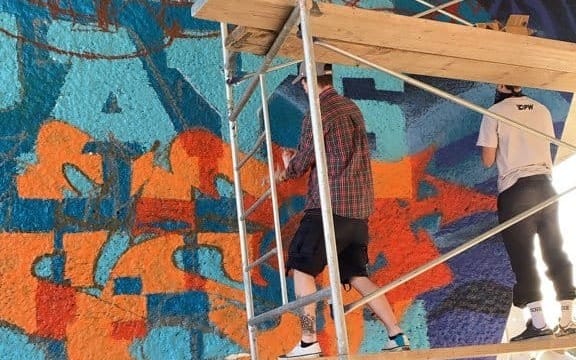 On voit les deux artistes de dos, sur un échafaudage, en train de travailler leur œuvre.