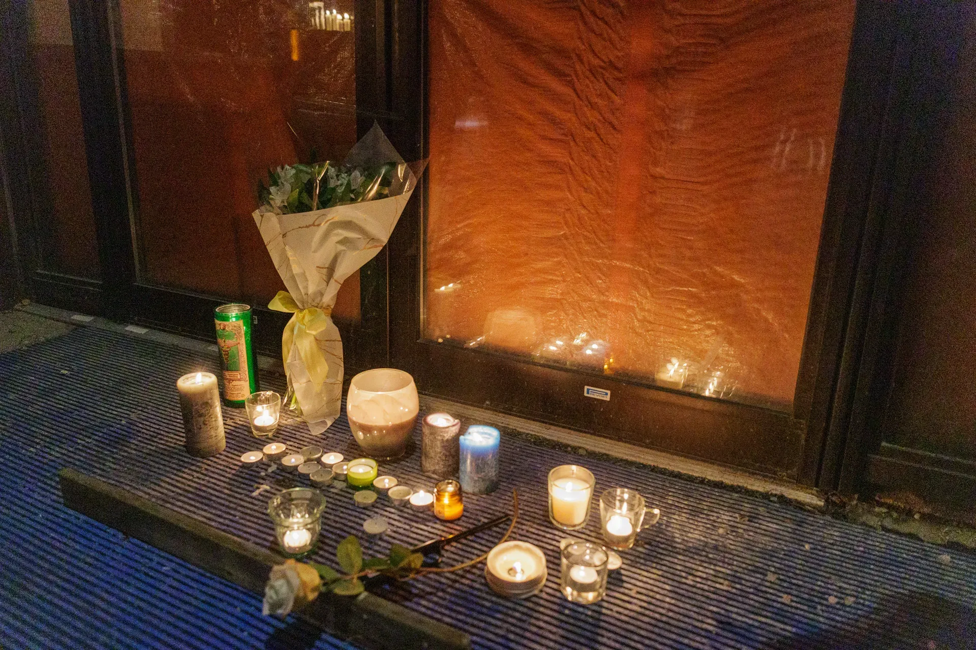 Un assortiment de petites bougies et de fleurs illumine le sol, devant l'entrée du dépanneur Fleur Bleue.