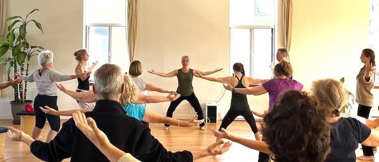 Un groupe de personnes s'étirent dans une salle lumineuse lors d'un cours de Qi Gong à l'école Fragments Libres.