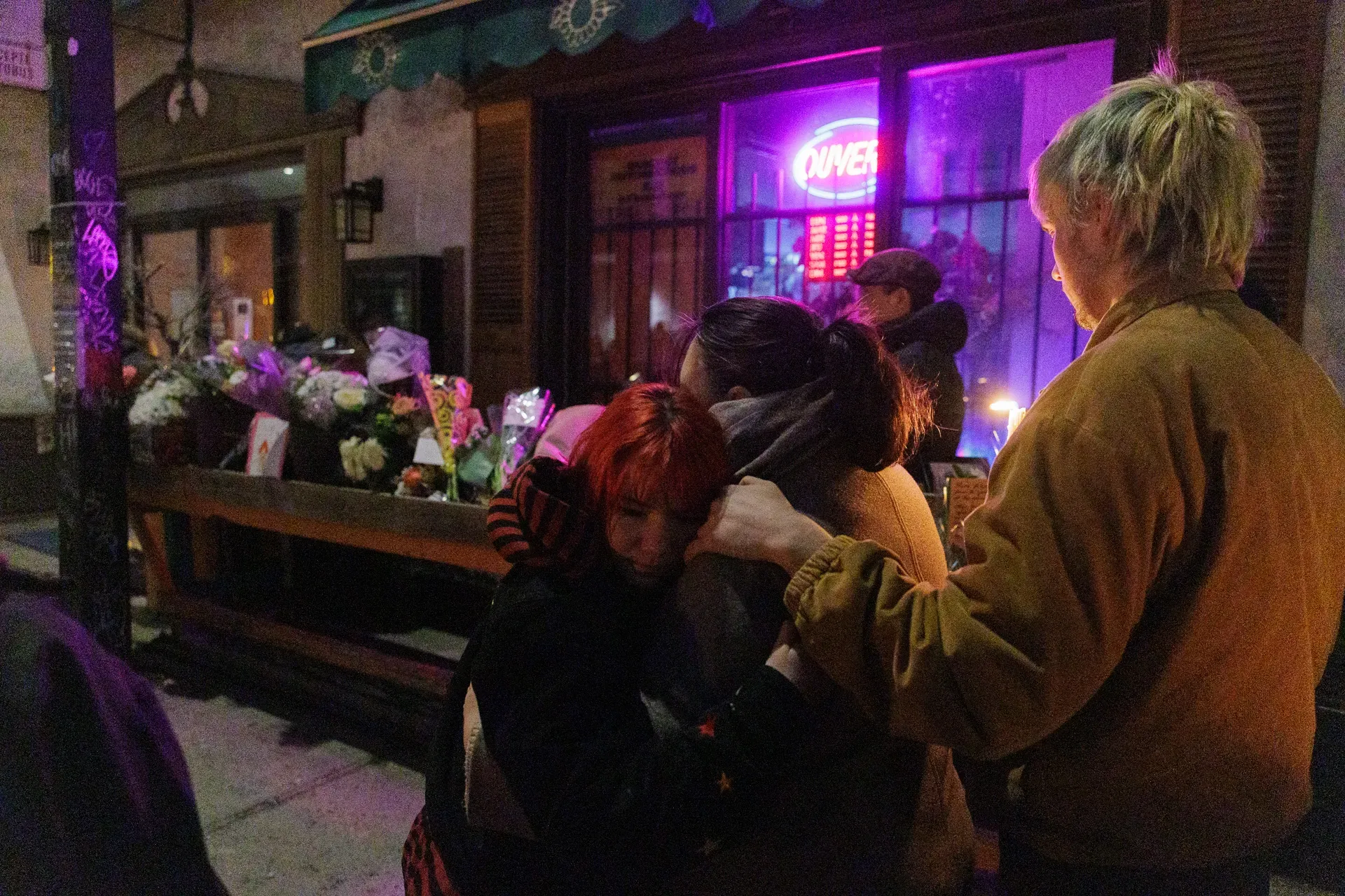 Deux femmes s'enlacent pendant la vigile. On voit les dizaines de bouquets de fleurs et la vitrine du commerce en arrière-plan.