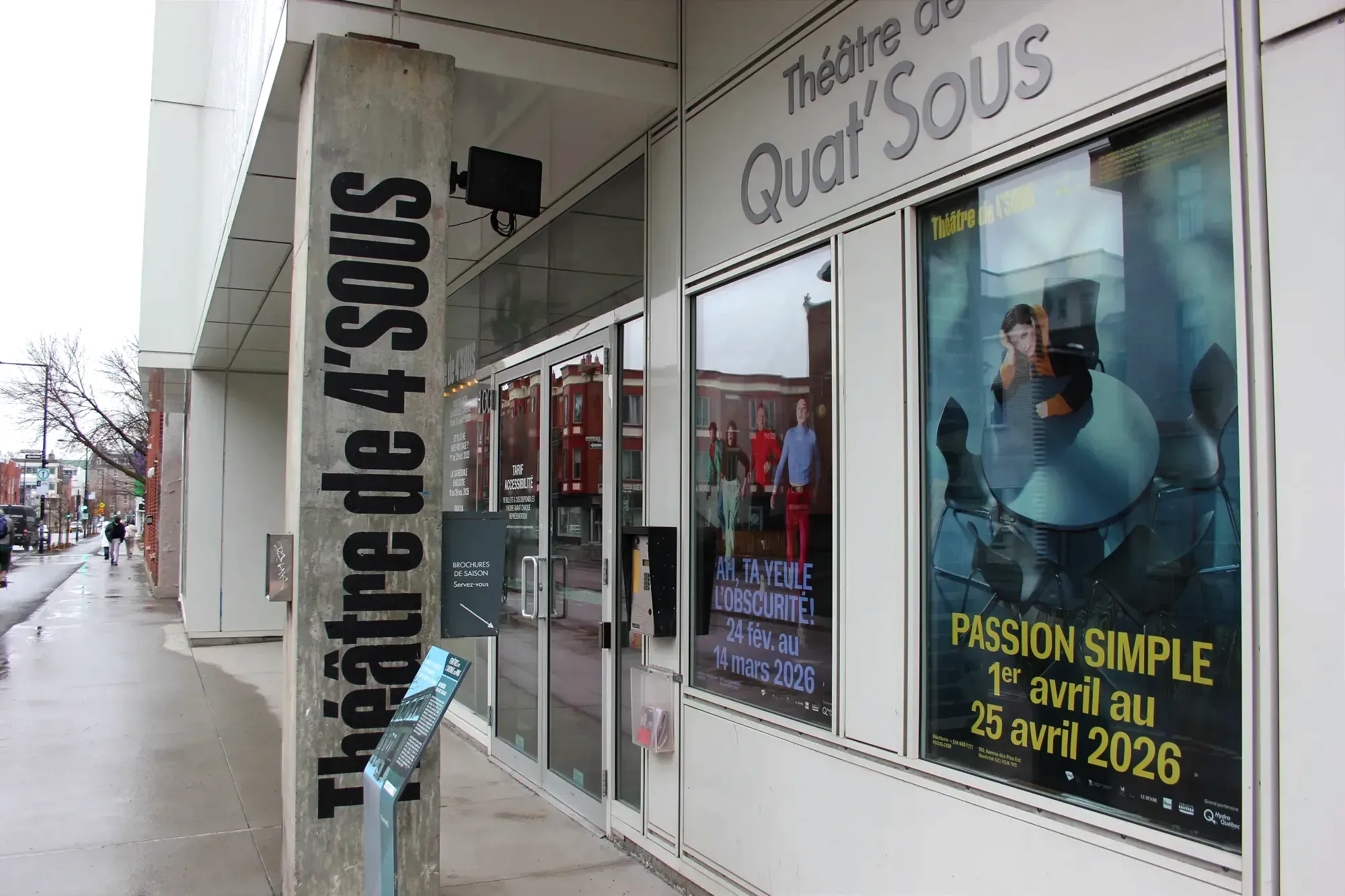 Vue extérieure des affiches près de l'entrée principale du théâtre, avec le trottoir de l'avenue des Pins dans le cadre.