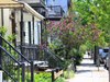 Un trottoir du Plateau-Mont-Royal illuminé en été. On y voit de balcons, un lilas en fleur et une femme qui marche au loin.