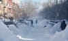 Des voitures sont largement cachées par des bancs de neige. On aperçoit plusieurs personnes et pelles dans la rue. 
