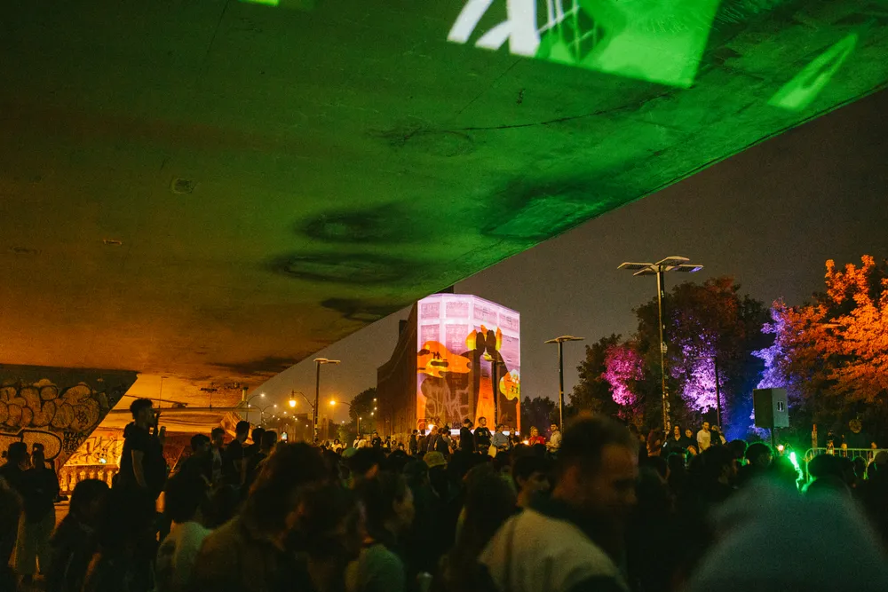 Une foule en-dessous d'un viaduc, le soir. Des projections sont visibles sur les structures environnantes.