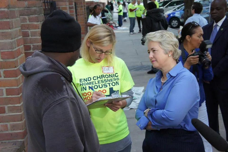 Une femme aux cheveux blonds, vêtue d'une chemise bleue et d'une jupe foncée (Annise Parker, l'ancienne mairesse de Houston) écoute attentivement un homme avec un chandail usé et une tuque.