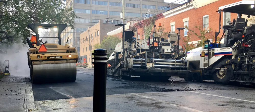 Un rouleau compresseur circule sur une petite rue du Plateau-Mont-Royal, entouré de camions d'asphaltage.